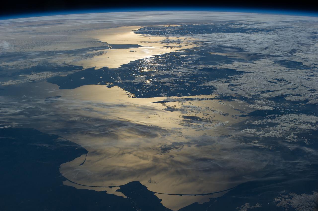 ISS040-E-012110 (15 June 2014) --- Coastlines of the southern Baltic Sea are featured in this image photographed by an Expedition 40 crew member on the International Space Station. The crew photographed this panorama looking aft (back along the orbital path) of the spacecraft as the sun was setting over the North Sea. Seen from the space station, the sun’s reflection point moves quickly across the landscape, momentarily lighting up water bodies. In this fleeting view the coast of southern Norway is outlined near the horizon. The strongest reflection of the sun highlights the narrow sea passage known as the Skagerrak—revealing the thin tip of Denmark. Numerous small lakes in southern Sweden appear at center, and scattered clouds cast complex shadows on the southern Baltic Sea. The sweeping curves of the sand spit on the Polish coast, and the long barrier islands on the Russian coast appear in the foreground at the edge of the sun’s reflection disc.