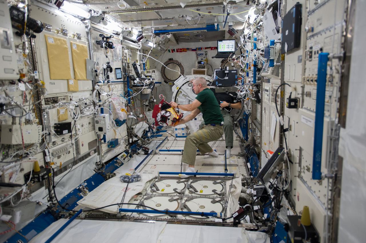 ISS040-E-015415 (18 June 2014) --- In the International Space Station's Kibo laboratory, NASA astronauts Steve Swanson, Expedition 40 commander; and Reid Wiseman (partially obscured), flight engineer, conduct test runs of the SPHERES-Slosh experiment, using the soccer-ball-sized, free-flying satellites known as Synchronized Position Hold, Engage, Reorient, Experimental Satellites, or SPHERES. The SPHERES-Slosh investigation uses small robotic satellites on the space station to examine how liquids move around inside containers in microgravity.