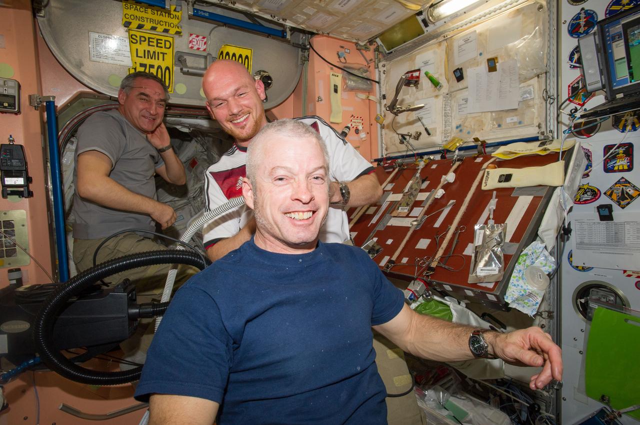 ISS040-E-020367 (26 June 2014) --- European Space Agency astronaut Alexander Gerst, Expedition 40 flight engineer, shaves the head of NASA astronaut Steve Swanson, commander, in the Unity node of the International Space Station. Gerst used hair clippers fashioned with a vacuum device to garner freshly cut hair. Russian cosmonaut Alexander Skvortsov, flight engineer, is visible in the background. A friendly World Cup Soccer-related agreement between the crew members representing the USA, and Gerst, a German citizen who represents the European Space Agency, called for the American losers of a German-USA match to have their heads shaved. The German team won the match 1-0.