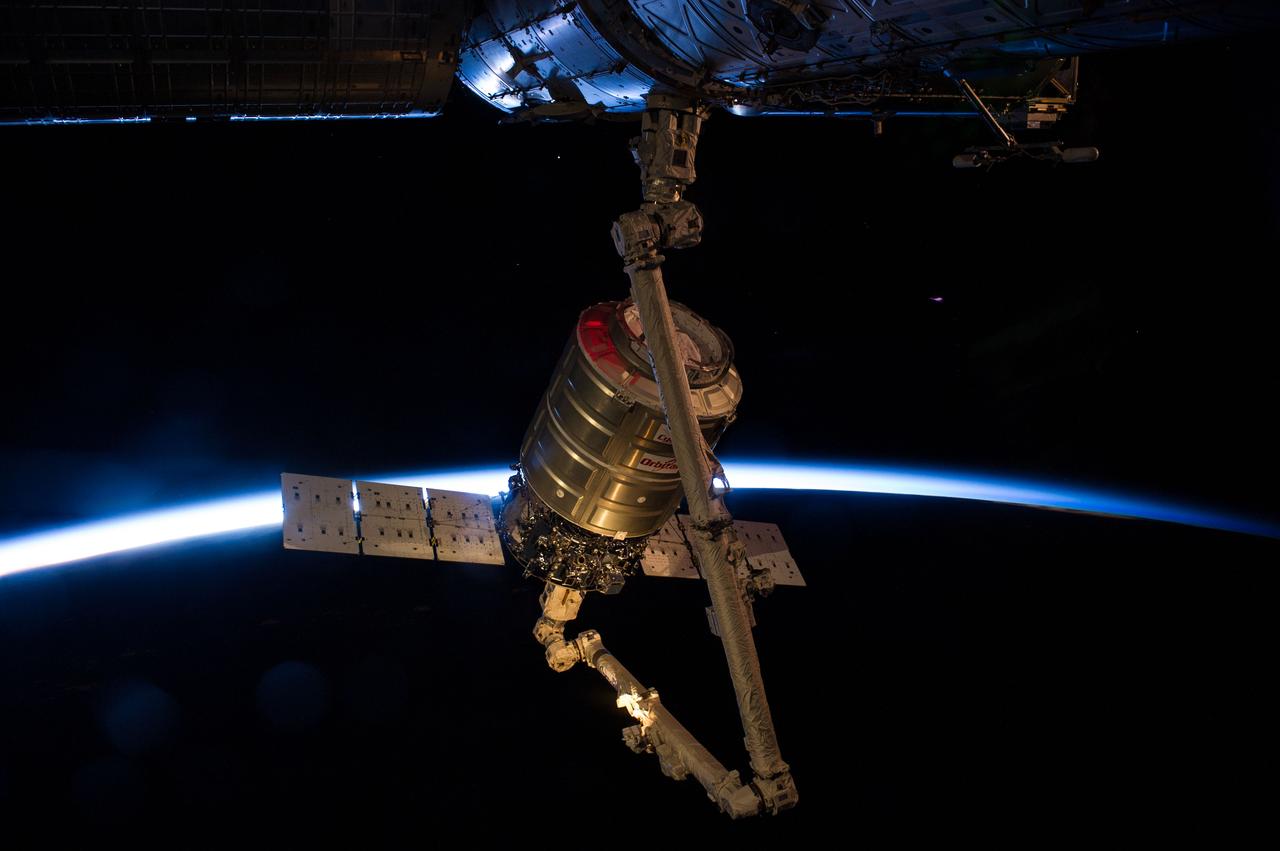 ISS040-E-063769 (16 July 2014) --- Intersecting the thin line of Earth?s atmosphere, the Orbital Sciences? Cygnus cargo craft attached to the end of the Canadarm2 robotic arm of the International Space Station is photographed by an Expedition 40 crew member after the two spacecraft converged at 6:36 a.m. (EDT) on July 16, 2014. The red color on Cygnus is a spectral effect from one of Cygnus? strobe lights against the fading light heading into an orbital sunset following grapple.