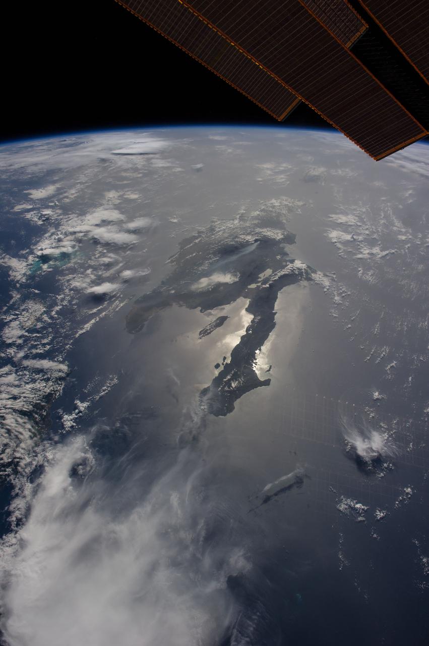 ISS040-E-080921 (26 June 2014) --- Dominican Republic and Haiti, Hispaniola, Caribbean are featured in this image photographed by an Expedition 40 crew member on the International Space Station. Looking east into a rising sun, the crew took this panorama of Hispaniola with the sun’s glint point illuminating the long western peninsula of Haiti. Several thunderheads throw shadows towards the camera (left). The plume from a very large wildfire stretches west (center). The Constanza Fire started in a national forest on the Dominican Republic growing to the extent that it threatened surrounding towns and prompting an International Disaster Charter activation, whereby requests for imagery were uplinked to the station crew as possible assistance to help firefighters on the ground. Hurricane Bertha tracked over the island a week later helping to douse the flames. The view looks hazy probably because of dust in the atmosphere. Dust blows across the Atlantic Ocean from Africa reaching the western hemisphere every month of the year. Despite the austere tone of the image, touches of color are blue waters of the Turks and Caicos Islands extending from under a large thundercloud (left) and the edge of a space station solar panel (top right).