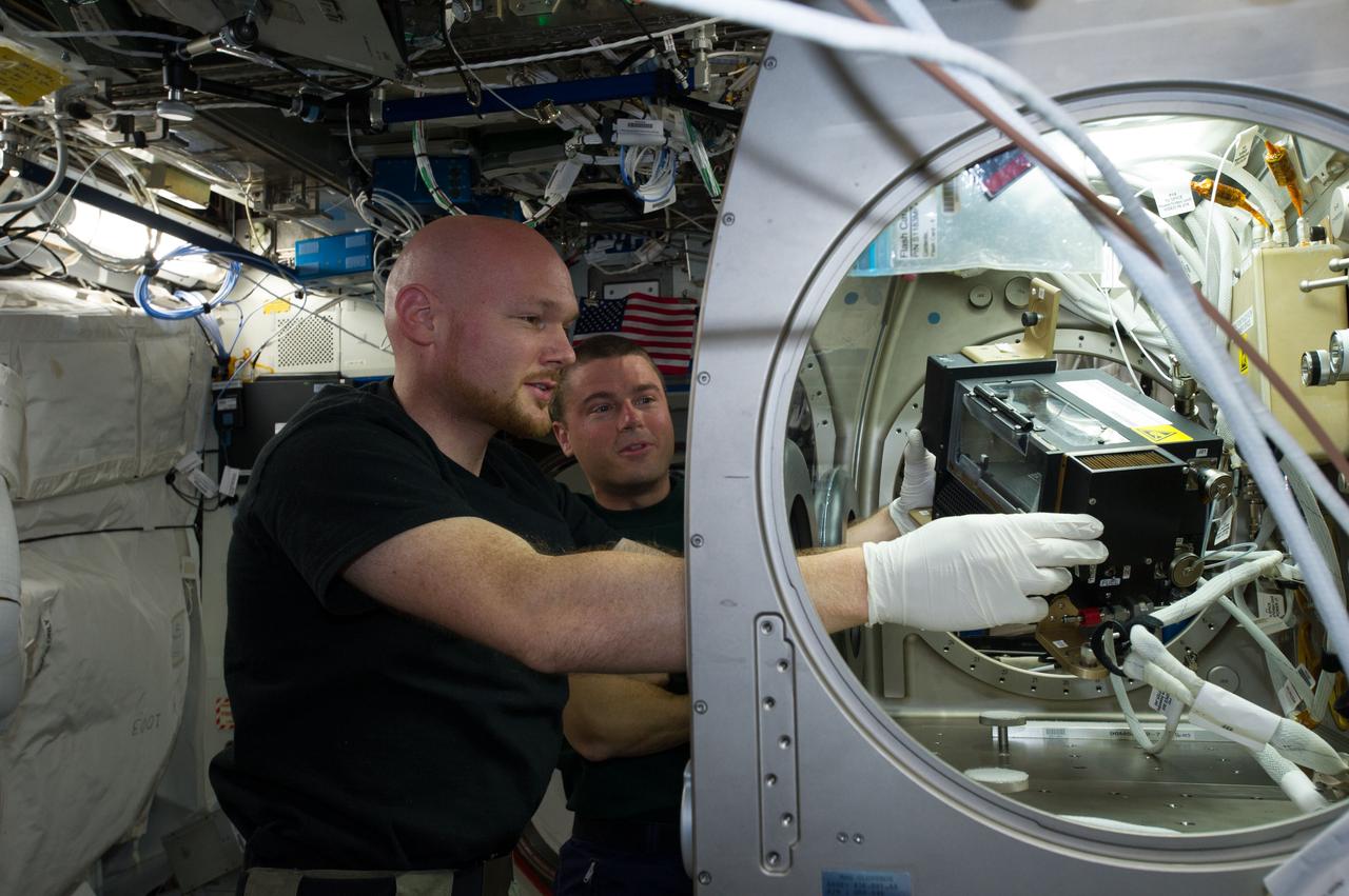 ISS040-E-088798 (5 Aug. 2014) --- European Space Agency astronaut Alexander Gerst, Expedition 40 flight engineer, removes hardware for the combustion experiment known as the Burning and Suppression of Solids (BASS-II) from the Microgravity Science Glovebox (MSG) in the Destiny laboratory of the International Space Station. The experiment seeks to provide insight on how flames burn in space compared to Earth which may provide fire safety benefits aboard future spacecraft. NASA astronaut Reid Wiseman, flight engineer, looks on.