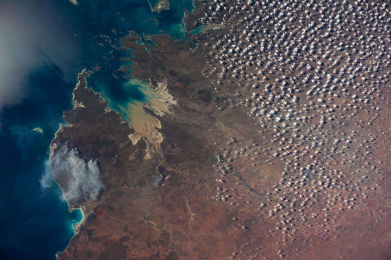 ISS040-E-089959 (8 Aug. 2014) --- King Sound on the northwest coast of Australia is featured in this image photographed by an Expedition 40 crew member on the International Space Station. The Fitzroy River, one of Australia's largest, empties into the Sound, a large gulf in Western Australia (approximately 120 kilometers long). King Sound has the highest tides in Australia, in the range of 11-12 meters, the second highest in the world after the Bay of Fundy on the east coast of North America. The strong brown smudge at the head of the Sound contrasts with the clearer blue water along the rest of the coast. This is mud stirred up by the tides and also supplied by the Fitzroy River. The bright reflection point of the sun obscures the blue water of the Indian Ocean (top left). Just to the west of the Sound, thick plumes of wildfire smoke, driven by northeast winds, obscure the coastline. A wide field of “popcorn cumulus” clouds (right) is a common effect of daily heating of the ground surface.