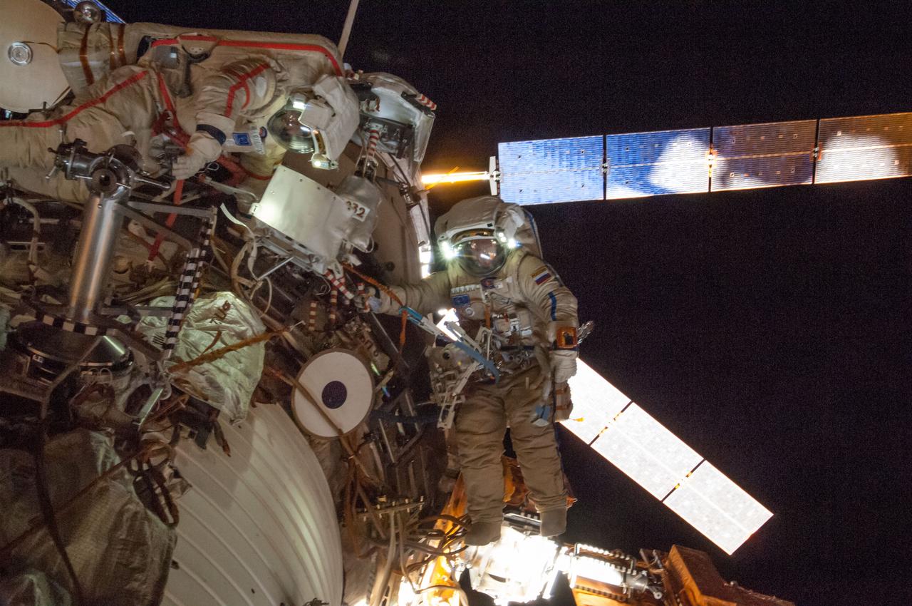 ISS040-E-099280 (18 Aug. 2014) --- Russian cosmonauts Alexander Skvortsov (left, red stripes) and Oleg Artemyev, both Expedition 40 flight engineers, attired in Russian Orlan spacesuits, participate in a session of extravehicular activity (EVA) in support of science and maintenance on the International Space Station. During the five-hour, 11-minute spacewalk, Artemyev and Skvortsov deployed a small science satellite, retrieved and installed experiment packages and inspected components on the exterior of the orbital laboratory. Solar array wings on the "Georges Lemaitre" Automated Transfer Vehicle-5 (ATV-5) docked to the Zvezda Service Module are visible in the background.