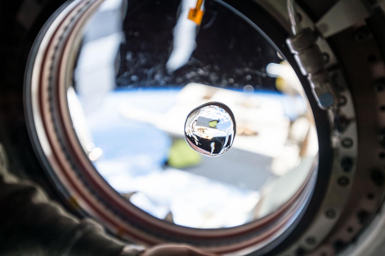 View of water bubble formed in front of the Japanese Experiment Module (JEM) window. The JEM Exposed Facility (JEF) is visible in the background through the window and reflected in the water. Scratches visible on the window.