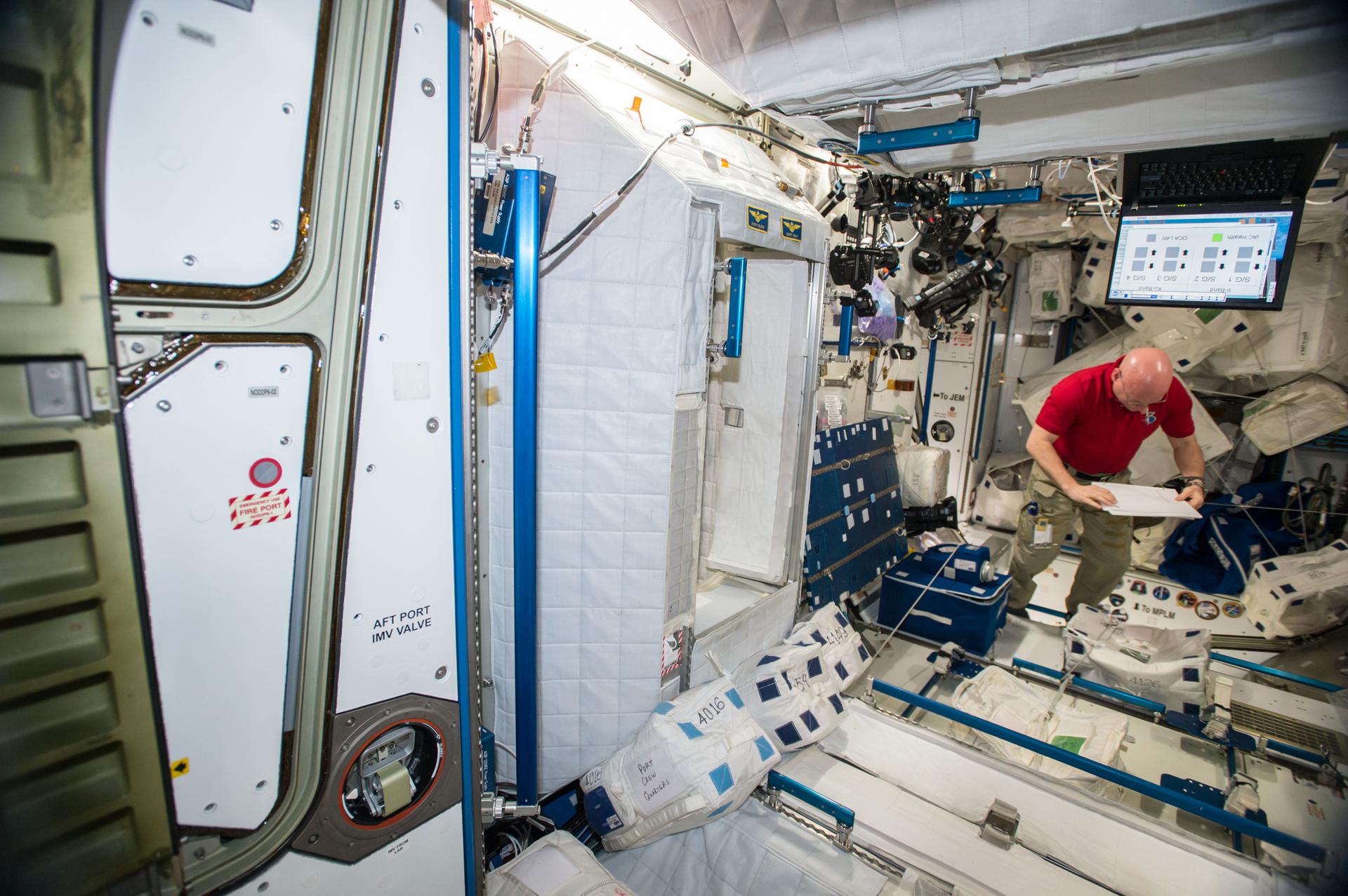Kelly, wearing a red shirt and khaki pants, is in the center of a space station module reading from a sheaf of papers. In front of him is a blue square container, one of the microbial samplers. A large laptop screen is above Kelly and multiple cameras are attached to the wall to his left.