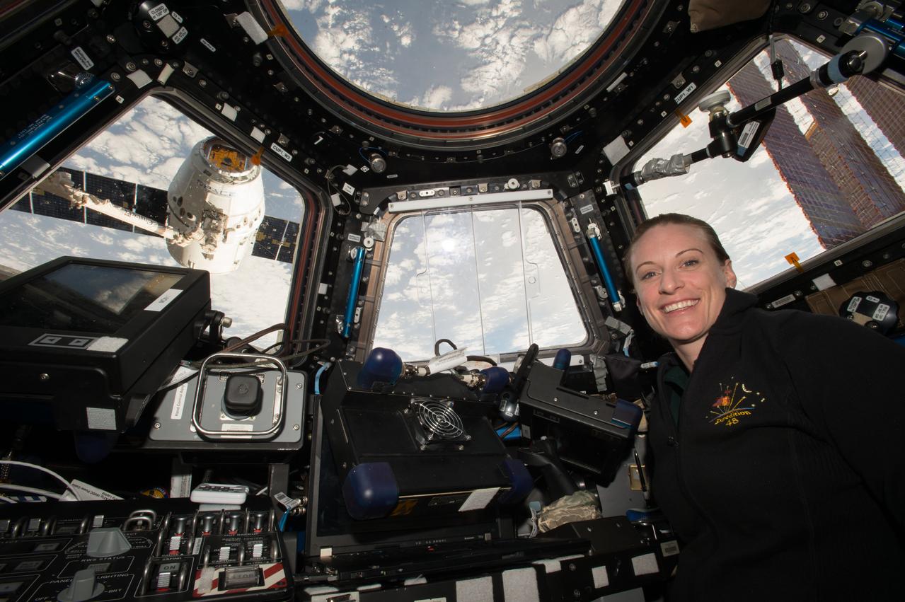 iss048e041862 (7/20/2016) --- NASA astronaut Kate Rubins poses for a photo in the Cupola module following SpaceX Dragon Commercial Resupply Services-9 (CRS-9) spacecraft grapple operations (OPS). The spacecraft is visible through Window 3.