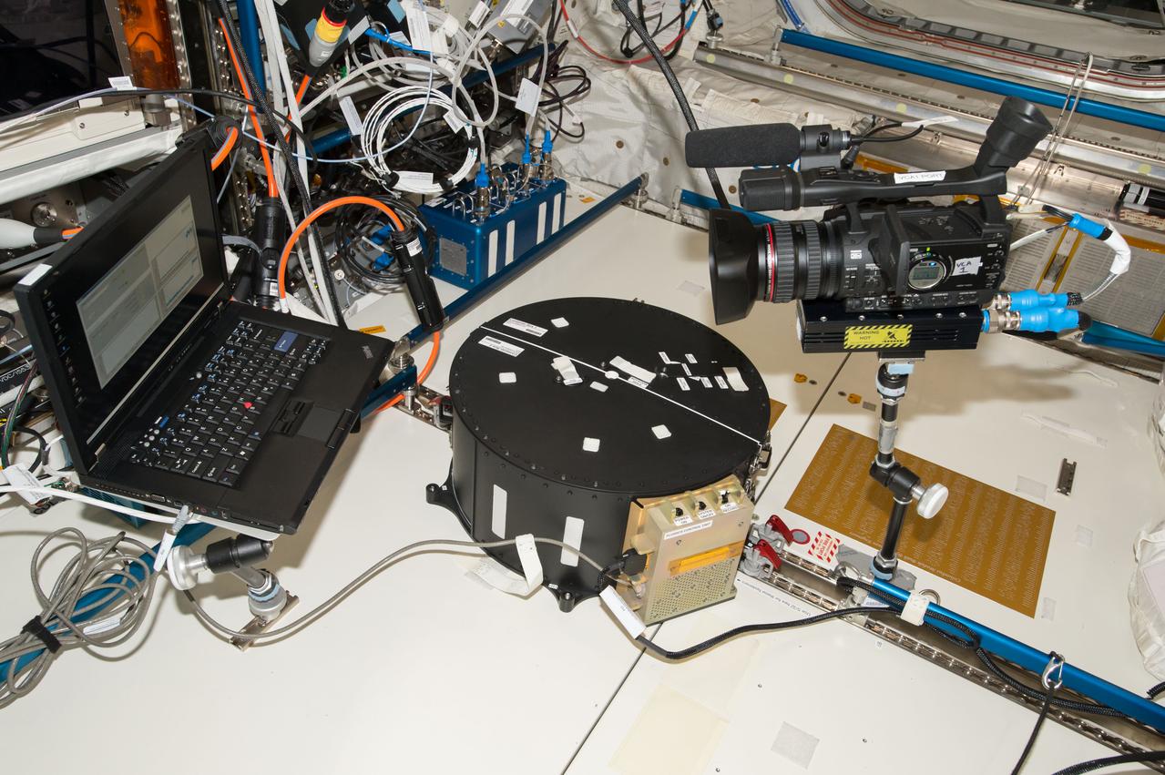 iss051e034000 (5/2/2017) --- A view taken to document hardware setup during the first run of the Fluid Dynamics in Space (FLUIDICS) experiment. The Container Box is attached to the Seat Track at COL1D1-D2, and a video camera records experiment data on the screen of a laptop computer. Image was taken in the Columbus European Laboratory. The FLUIDICS investigation evaluates the Center of Mass (CoM) position regarding a temperature gradient on a representation of a fuel tank. The observation of capillary wave turbulence on the surface of a fluid layer in a low-gravity environment can provide insights into measuring the existing volume in a sphere.