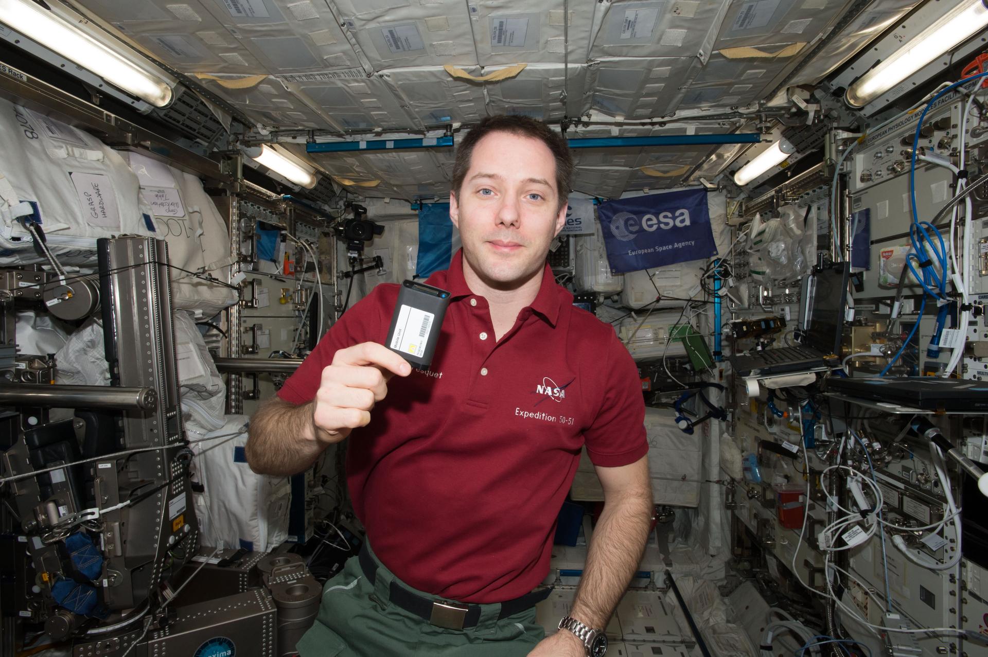 Pesquet, wearing a red polo, green pants, and a black belt, holds a rectangular black device smaller than a cell phone in his right hand. An ESA flag is visible over his left shoulder, equipment and storage bags to his right, and several laptops and cables to his left.