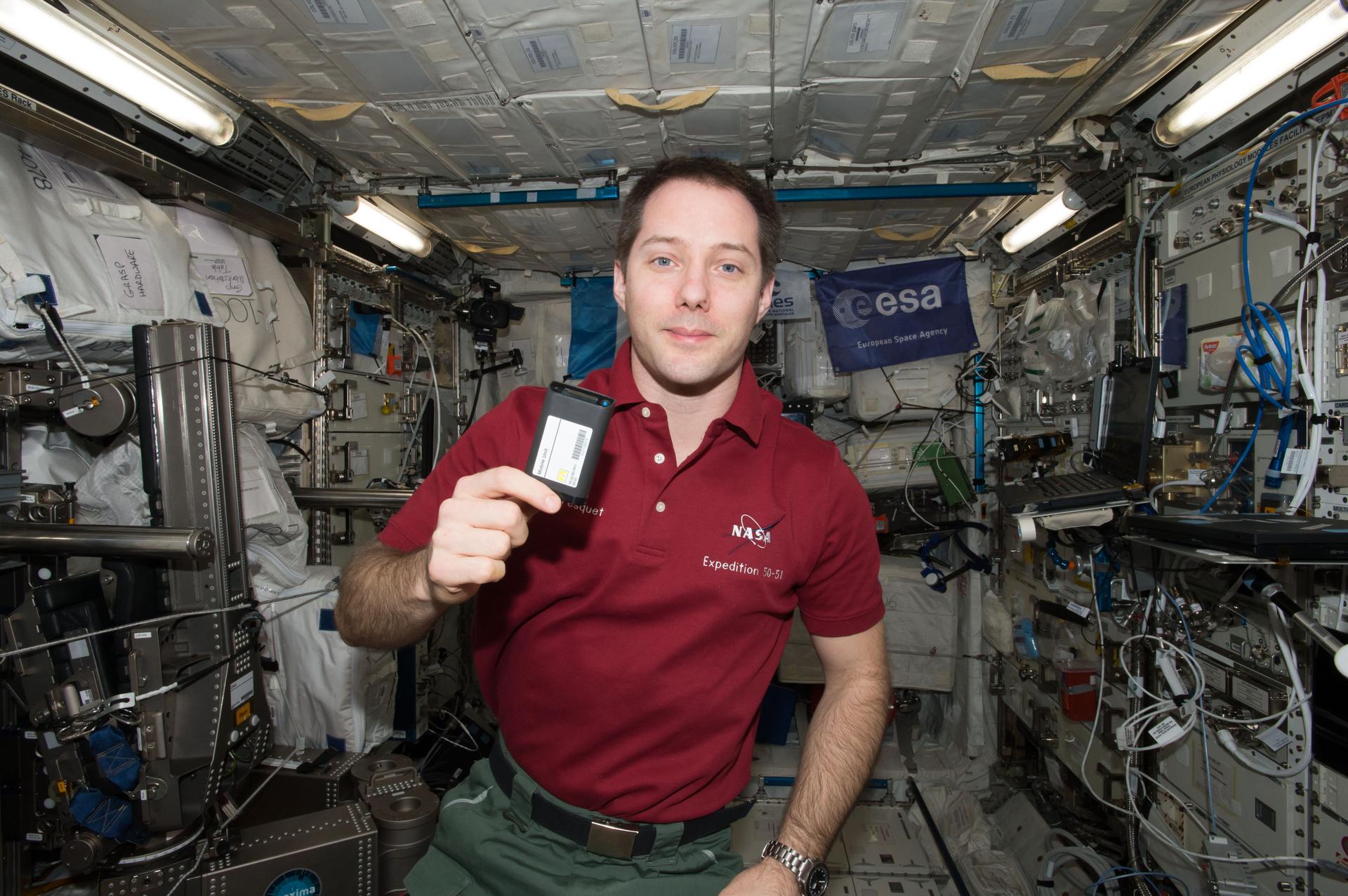 Pesquet, wearing a red polo shirt and green pants, holds a small, black rectangular device the size of a pack of cards. The device has a large white label on the front and on its end, a gold sensor strip and a blue light.
