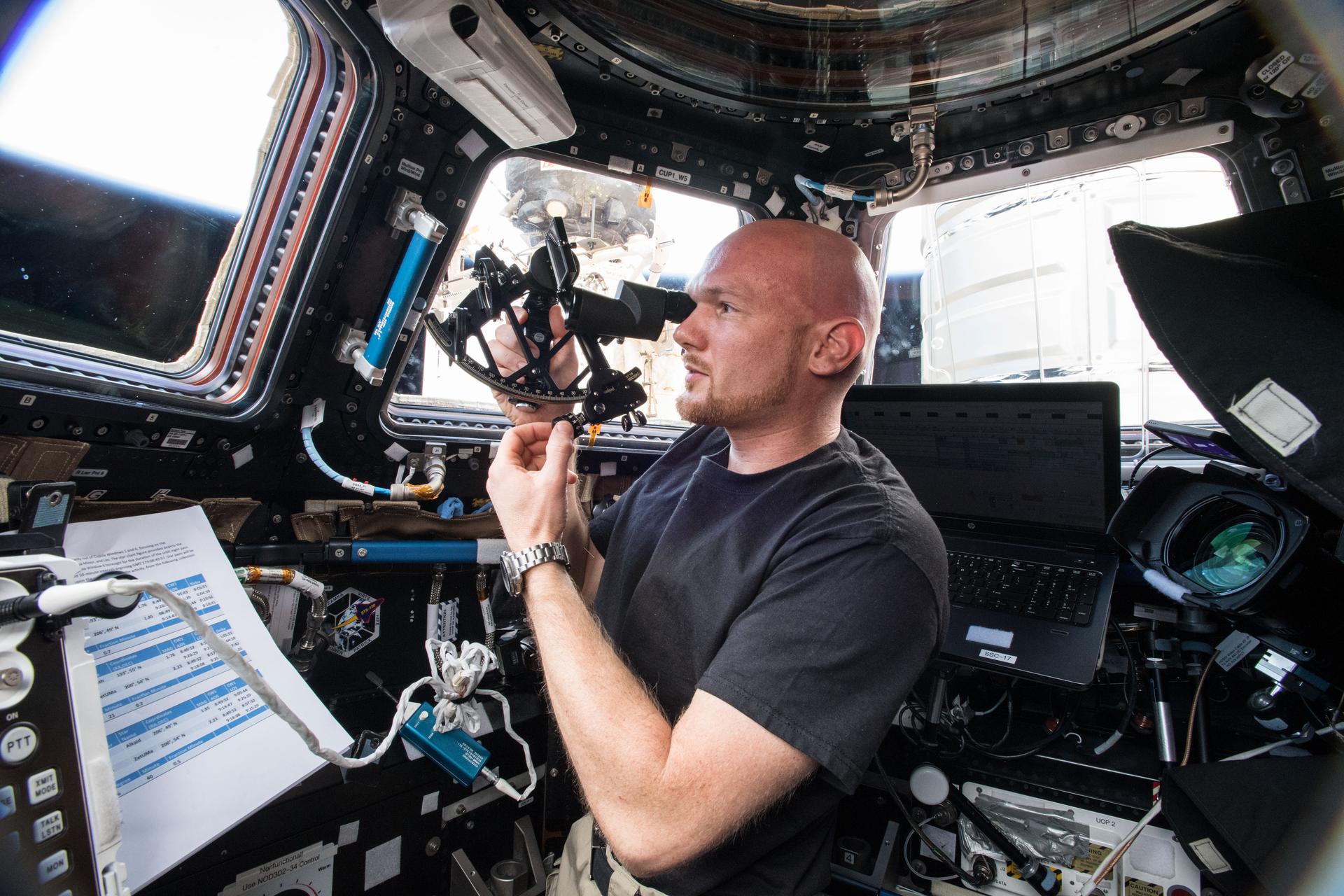 Gerst, wearing a short-sleeved black t-shirt and a watch on his left wrist, holds the sextant up to his right eye as he faces a window in the cupola. The black device has a sighting eye piece and a curved positioning piece across the bottom that Gerst is adjusting with his left hand. There is a laptop behind him and a sheet of instructions in front of him.