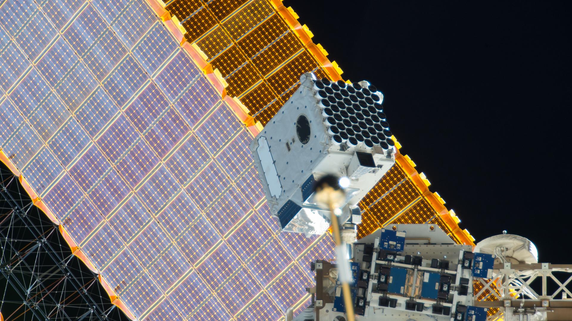 A large white box covered on one side in multiple circular black sensors points out into space. One of the station’s large solar panels is visible behind it against the blackness.