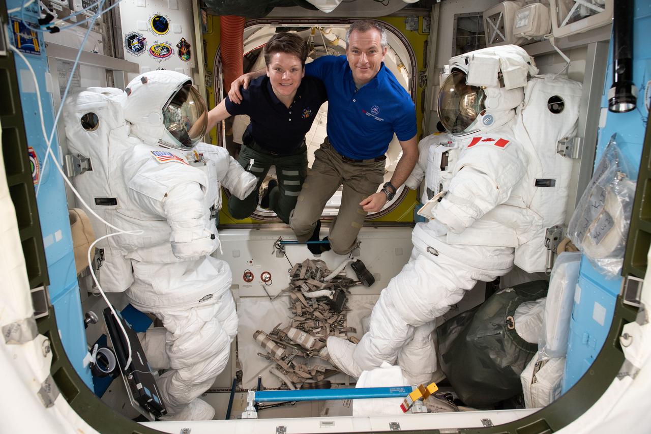 iss058e015358 (Feb. 17, 2019) --- Astronauts (from left) Anne McClain and David Saint-Jacques are pictured in between a pair of spacesuits that are stowed and serviced inside the Quest airlock where U.S. spacewalks are staged.
