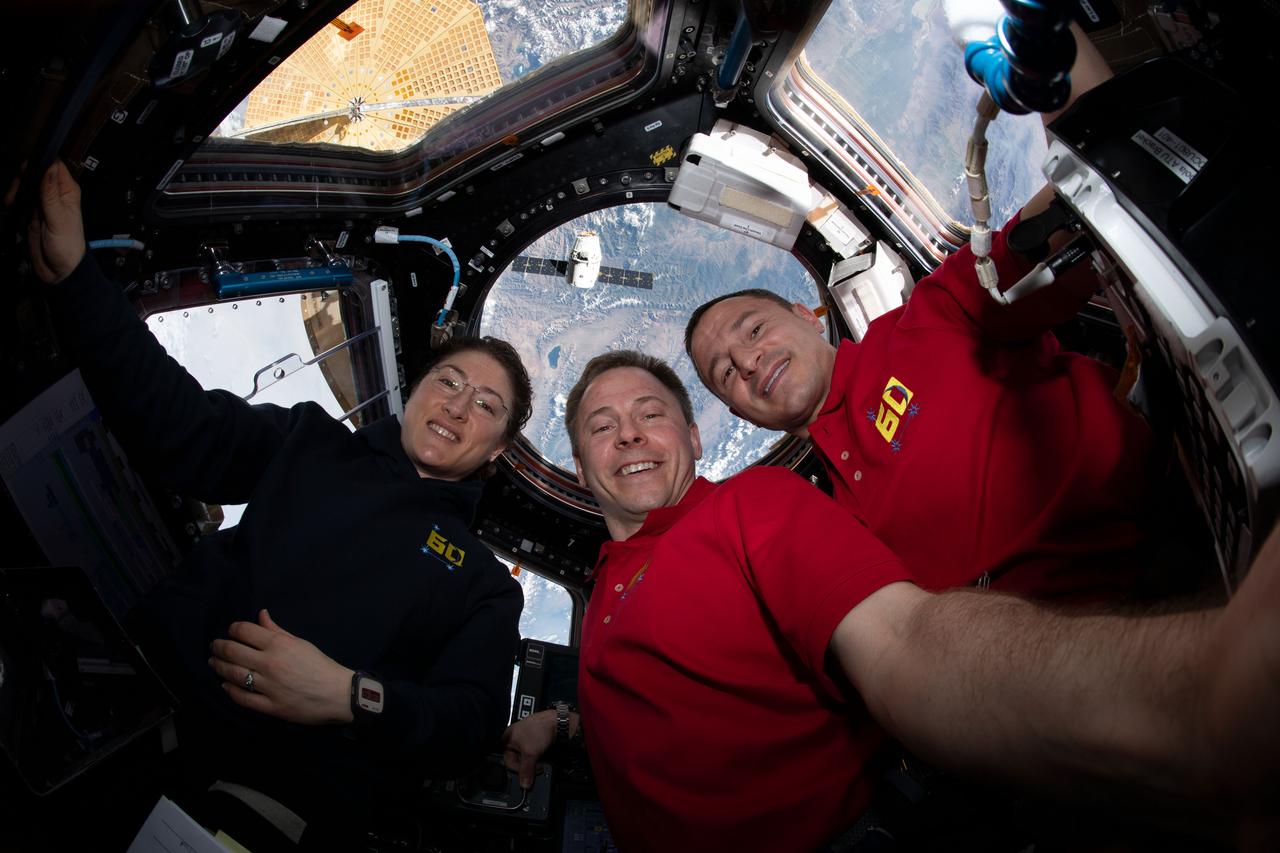 iss060e015993 (July 27, 2019) --- NASA astronauts (from left) Christina Koch, Nick Hague and Andrew Morgan gather for a portrait inside the International Space Station's "window to the world," the seven-windowed cupola. Behind the trio, the SpaceX Dragon cargo craft is approaching the orbiting lab for a robotic capture as both spacecraft were orbiting 261 miles above Tajikistan.