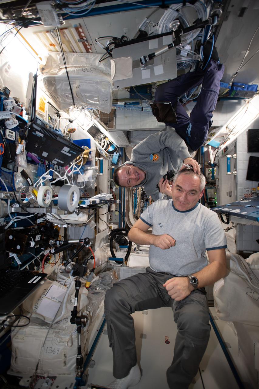 iss061e005740 (Oct. 13, 2019) --- NASA astronaut Andrew Morgan (right) took a break from his engineering and science duties aboard the International Space Station to trim the hair of Roscosmos cosmonaut Alexander Skvortsov. A vacuum is attached to the clippers that draws the loose hair in to keep the cabin atmosphere clean.