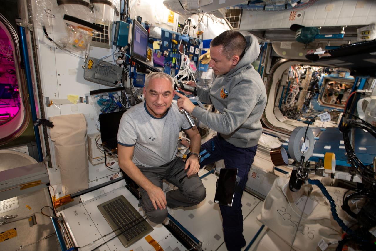 iss061e005754 (Oct. 13, 2019) --- NASA astronaut Andrew Morgan (right) took a break from his engineering and science duties aboard the International Space Station to trim the hair of Roscosmos cosmonaut Alexander Skvortsov. A vacuum is attached to the clippers that draws the loose hair in to keep the cabin atmosphere clean.