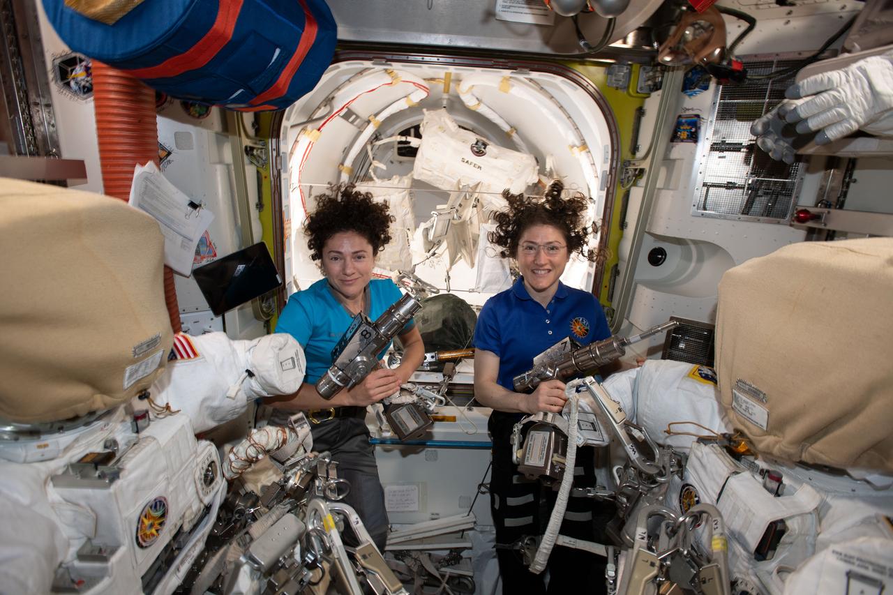 iss061e006498 (Oct. 15, 2019) --- NASA astronauts Jessica Meir (left) and Christina Koch are inside the Quest airlock preparing the U.S. spacesuits and tools they will use on their first spacewalk together. The Expedition 61 flight engineers are holding the pistol grip tools they will use to swap out a failed power controller, also known as a battery charge-discharge unit, that regulates the charge to batteries that collect and distribute power to the International Space Station.