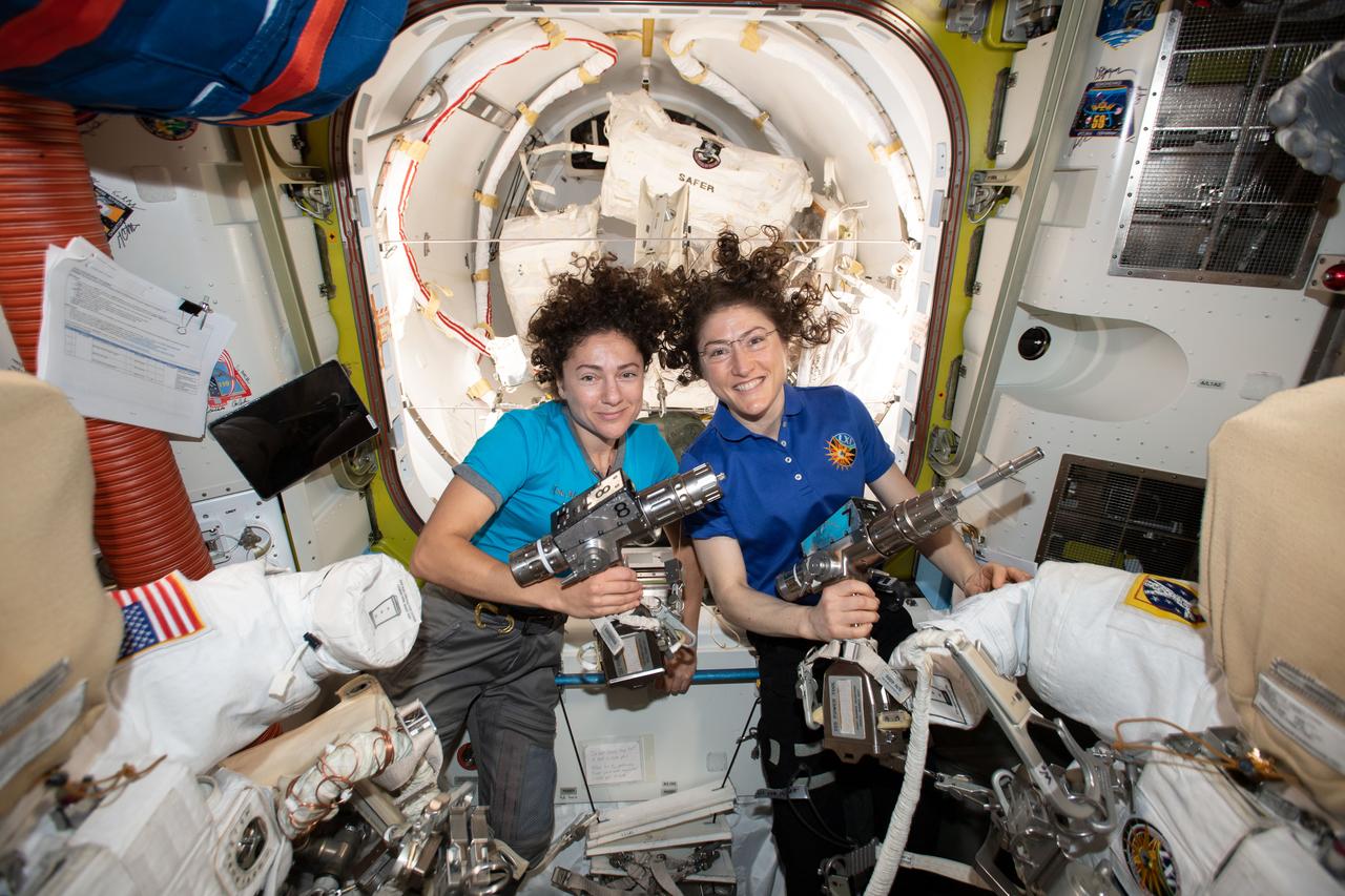 iss061e006501 (Oct. 15, 2019) --- NASA astronauts Jessica Meir (left) and Christina Koch are inside the Quest airlock preparing the U.S. spacesuits and tools they will use on their first spacewalk together. The Expedition 61 flight engineers are holding the pistol grip tools they will use to swap out a failed power controller, also known as a battery charge-discharge unit, that regulates the charge to batteries that collect and distribute power to the International Space Station.