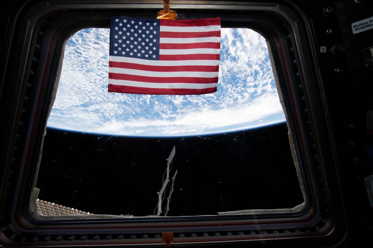 A United States flag floats in front of a window in the Cupola module.