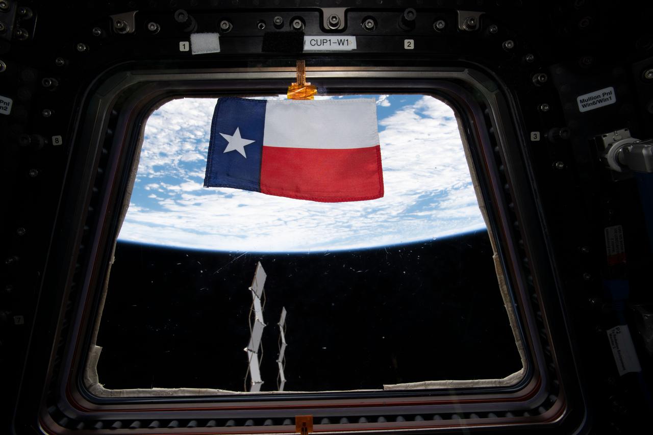 A state flag of Texas floats in front of a window in the Cupola module.