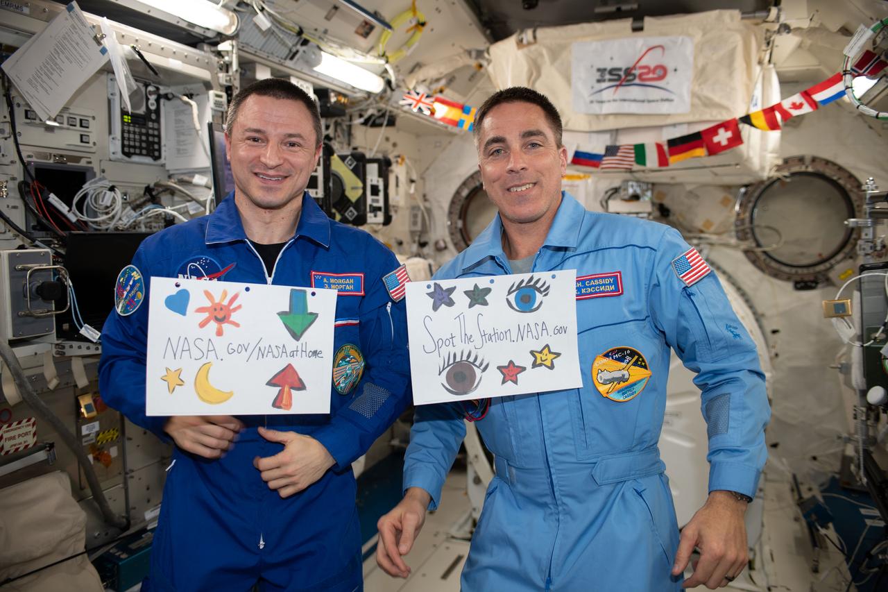 iss062e147663 (4/12/2020) --- A view taken during candid shots in Japanese Experiment Module (JEM) aboard the International Space Station (ISS) during PAO event. Astronaut Drew Morgan, in his blue flight suit, participates in the event. He is holding a sign that reads NASA.gov/NASAatHome. He is joined by astronaut Christopher Cassidy holding a sign that reads Spot the station.NASA.gov.