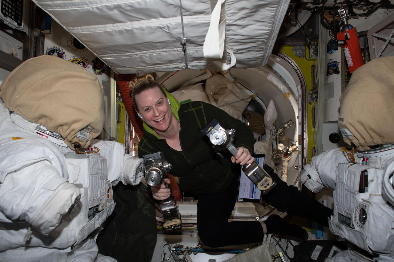 iss064e013381 (Dec. 18, 2020) --- NASA astronaut and Expedition 64 Flight Engineer Kate Rubins is pictured inside the U.S. Quest airlock carrying a pair of pistol grip tools used for maintenance work during spacewalks. Rubins is flanked by a pair of U.S. spacesuits with the arm segments unattached and the helmets covered for protection.