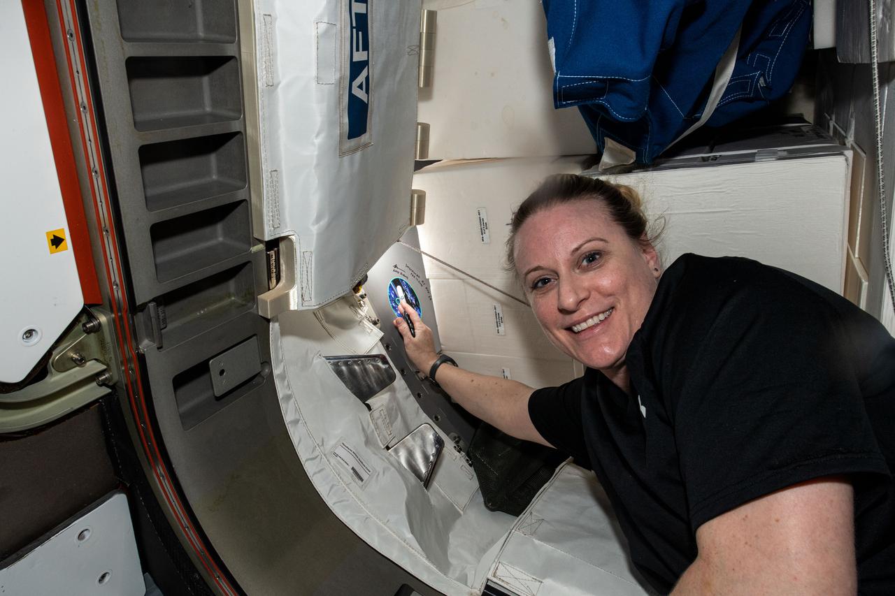 iss064e031800 (Feb. 11,2021) --- NASA astronaut and Expedition 64 Flight Engineer Kate Rubins signs her name near her mission's insignia sticker in the vestibule between the Tranquility module and the NanoRacks Bishop airlock.