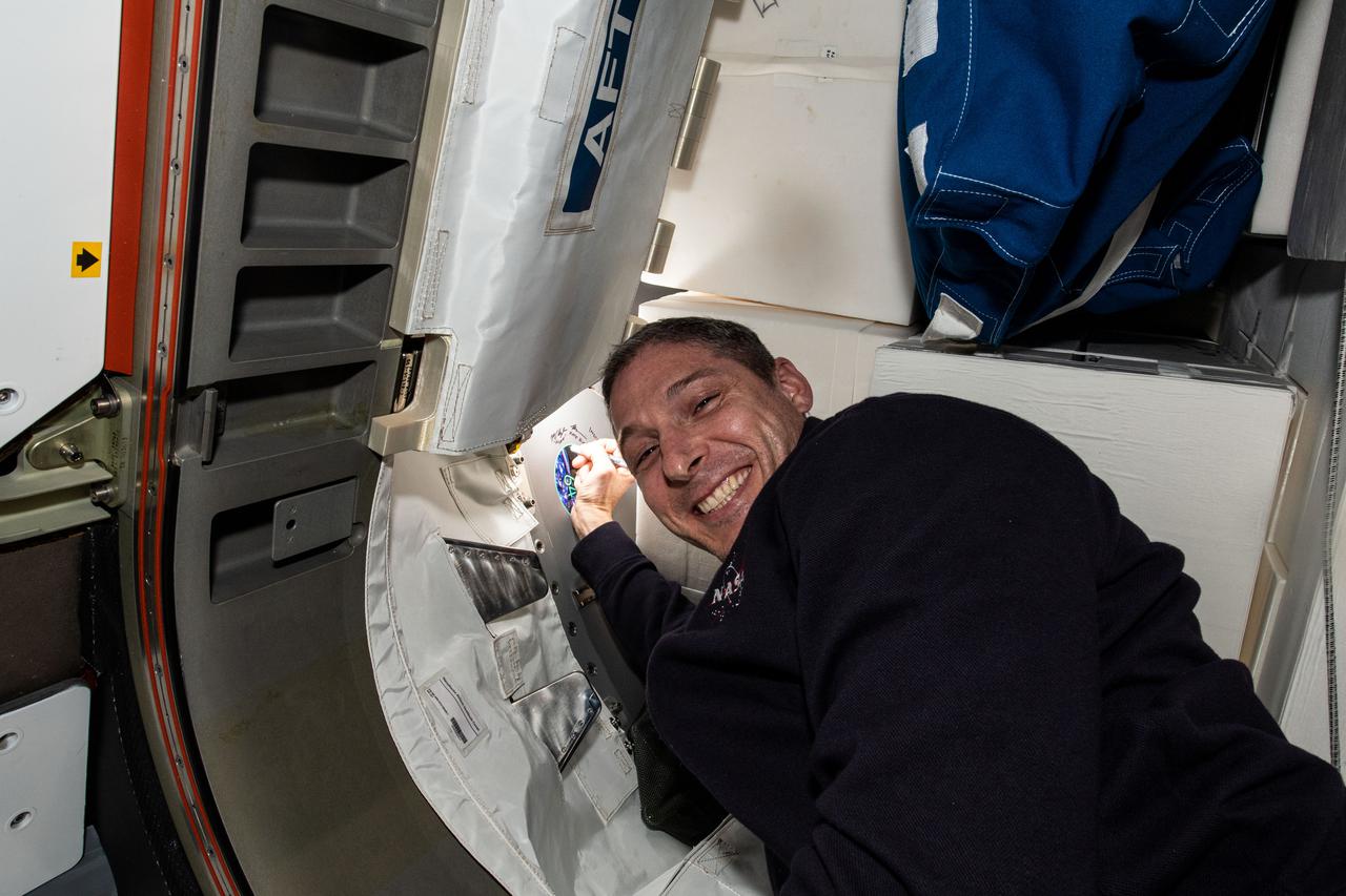 iss064e031801 (Feb. 11,2021) --- NASA astronaut and Expedition 64 Flight Engineer Michael Hopkins signs his name near his mission's insignia sticker in the vestibule between the Tranquility module and the NanoRacks Bishop airlock.
