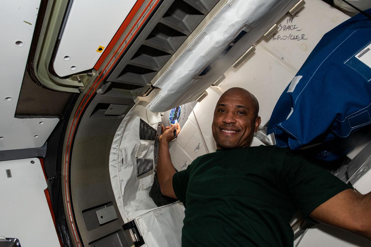 iss064e031803 (Feb. 11,2021) --- NASA astronaut and Expedition 64 Flight Engineer Victor Glover signs his name near his mission's insignia sticker in the vestibule between the Tranquility module and the NanoRacks Bishop airlock.