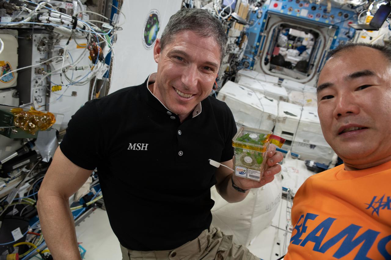 iss064e043776 (March 16, 2021) --- Expedition 64 Flight Engineers Michael Hopkins of NASA and Soichi Noguchi of JAXA (Japan Aerospace Exploration Agency) are pictured inside the International Space Station's Kibo laboratory module. Hopkins is showing a plant chamber containing  plants growing for the Asian Herb in Space (AHiS) study. The space botany investigation is exploring ways to cultivate fast-growing plants used for traditional medicine and food flavoring.