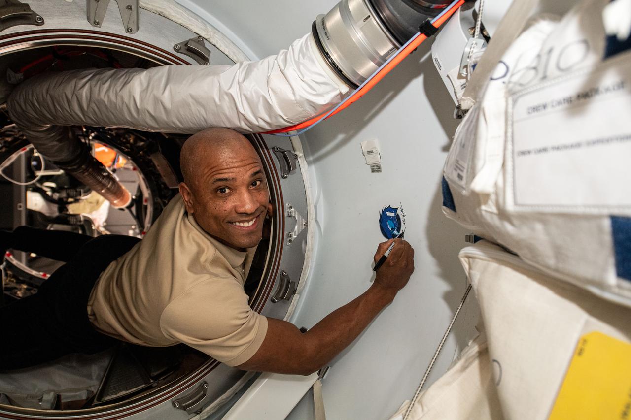 iss064e052200 (April 4, 2021) --- SpaceX Crew-1 Pilot Victor Glover of NASA signs his name next to the mission insignia affixed to the vestibule between the Crew Dragon spacecraft and the Harmony module's forward international docking adapter.