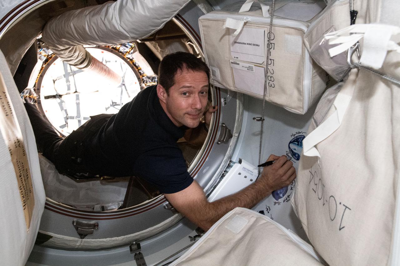 iss065e162503 (July 6, 2021) --- Expedition 65 Flight Engineer Thomas Pesquet of ESA (European Space Agency) signs his name next to the SpaceX CRS-22 cargo mission sticker attached to the vestibule between the SpaceX Cargo Dragon resupply ship and the Harmony module's space-facing international docking adapter.