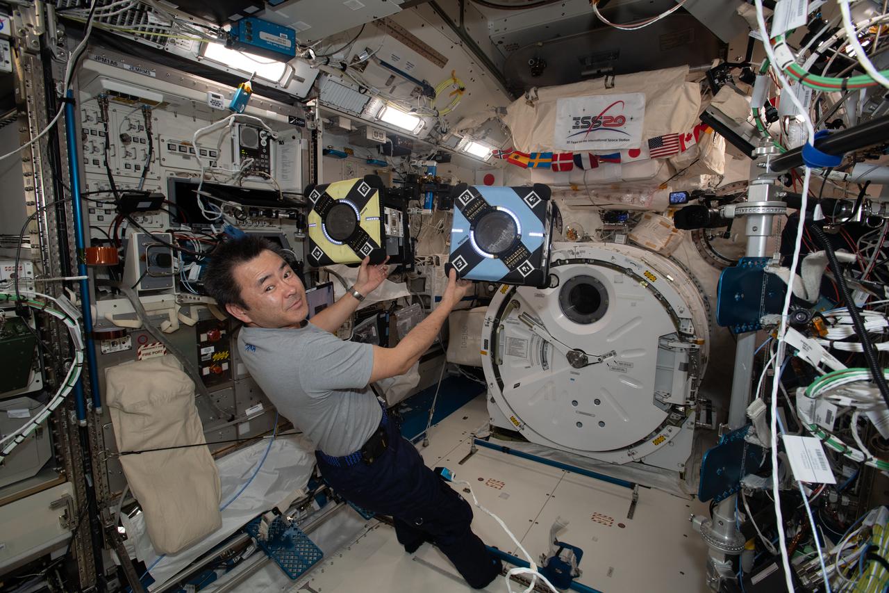 iss065e319330 (Aug. 26, 2021) --- Expedition 65 Commander Akihiko Hoshide of the Japan Aerospace Exploration Agency checks out a pair of Astrobee robotic free-flyers. The toaster-sized robotic assistants are being tested for their ability to autonomously conduct routine orbital chores, monitor station activities and coordinate maneuvers for future satellite repair techniques.