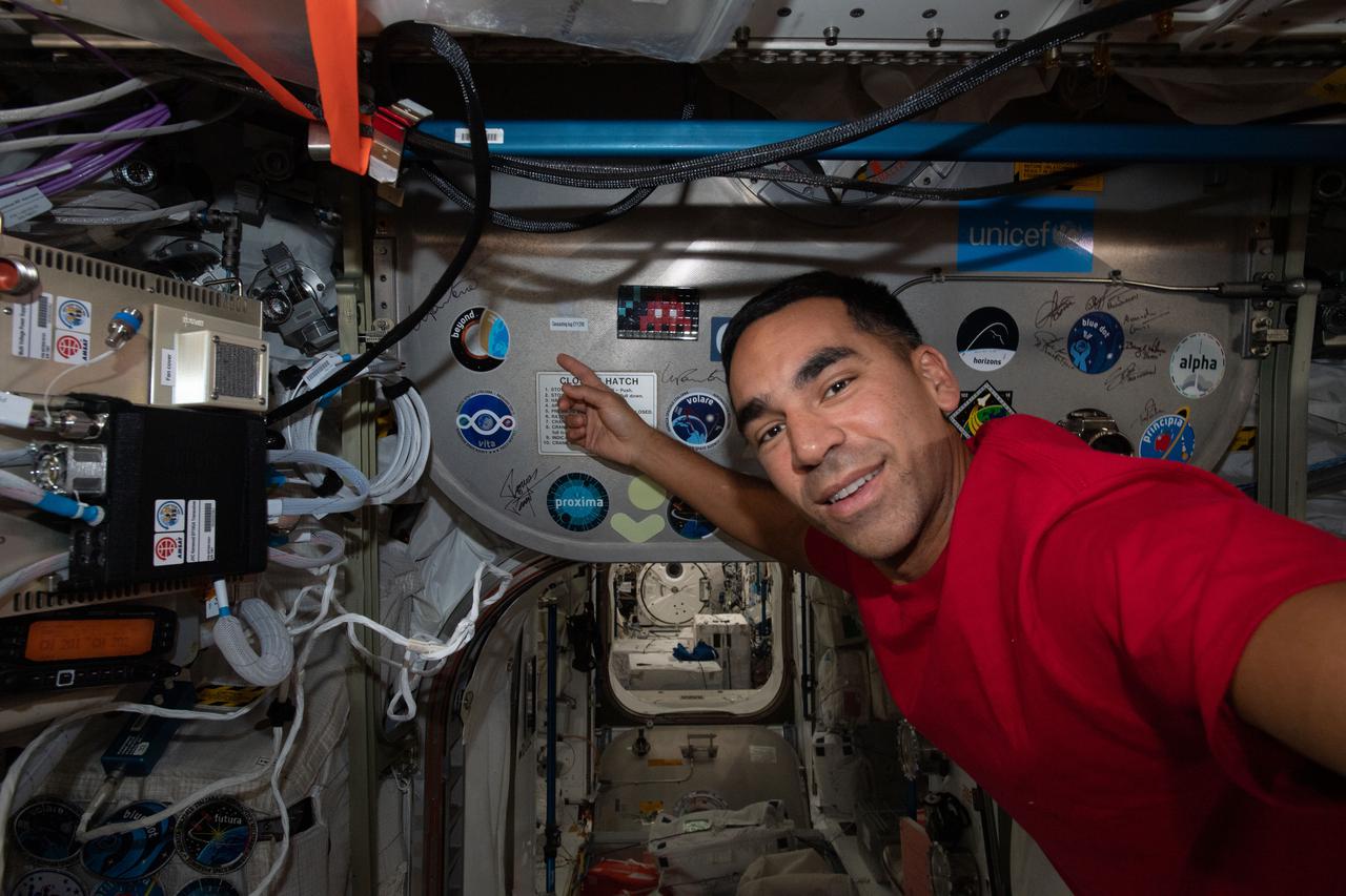 iss066e086630 (Dec. 5, 2021) --- NASA astronaut and Expedition 66 Flight Engineer Raja Chari is pictured inside the Columbus laboratory module pointing to the "Beyond" mission insignia representing ESA (European Space Agency) astronaut Luca Parmitano's stay aboard the International Space Station.