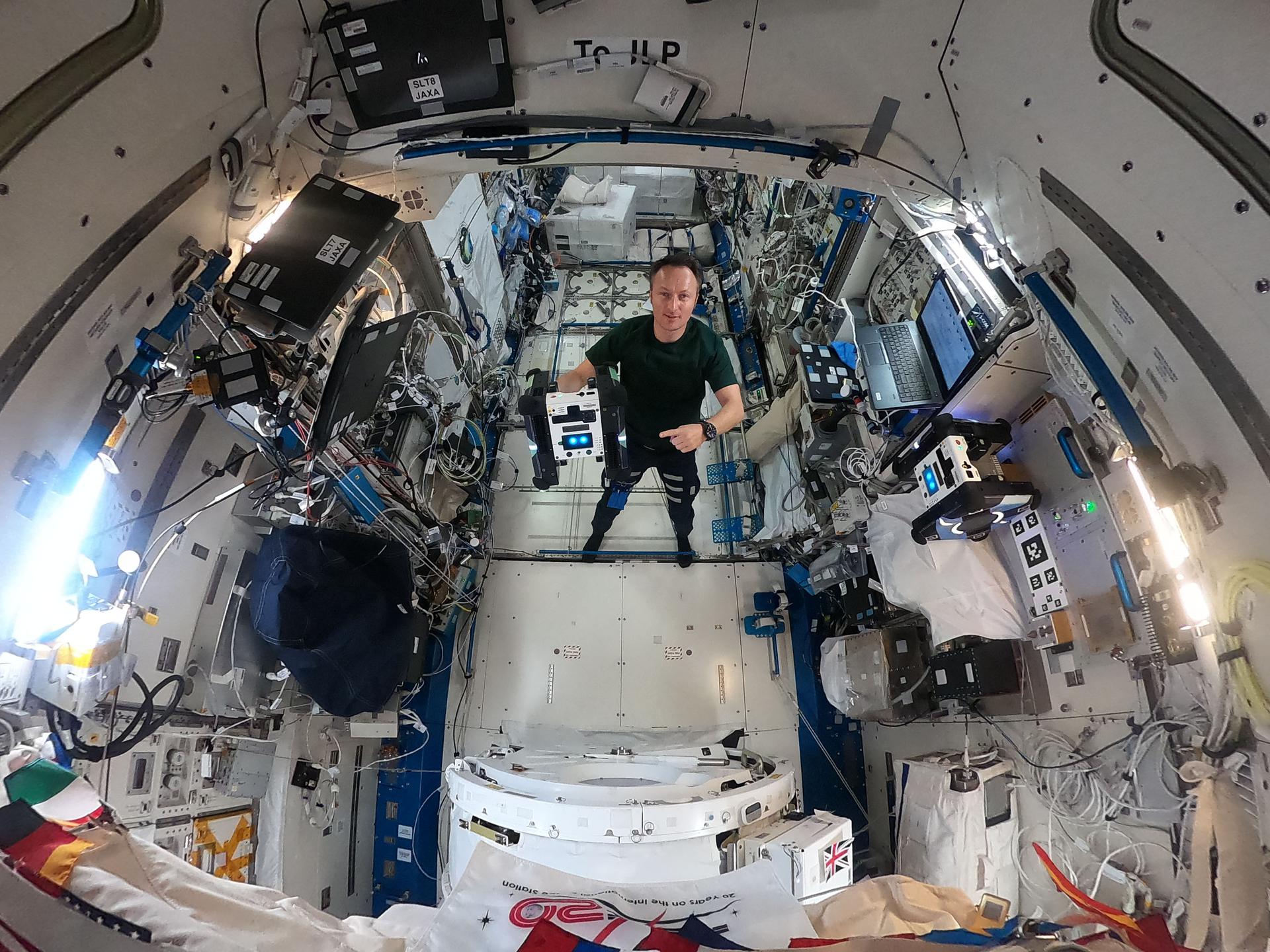 Maurer stands in the middle of this circular fisheye image, facing the camera, wearing a green t-shirt and black pants. In his right hand, he holds a cube-shaped Astrobee, which has black sides and a white panel facing the camera with two bright blue lights that look sort of like eyes. Maurer is pointing at the Astrobee with his left hand.