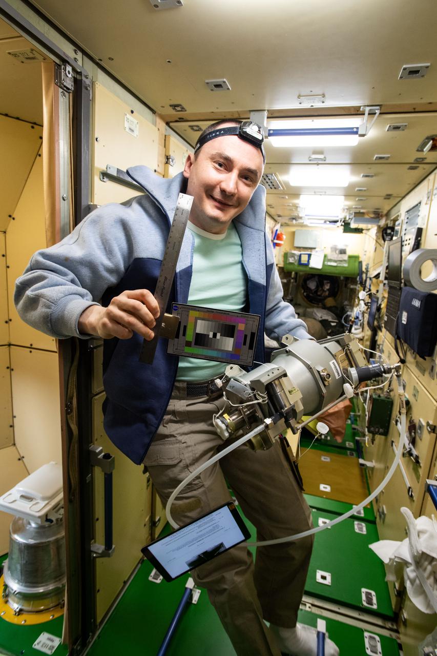 iss066e129780 (Jan. 28, 2022) --- Roscosmos cosmonaut and Expedition 66 Flight Engineer Pyotr Dubrov poses with a ruler and color chart while working inside the Nauka multipurpose laboratory module. The ruler and chart are used for the SQuARES study looking at how crew members use different objects and spaces over time. This investigation may provide information that helps improve the design of future space craft and habitats. At bottom left, is the Nauka module's toilet.