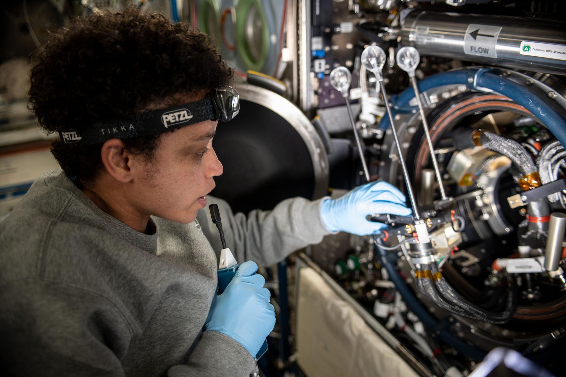 A female astronaut wearing blue gloves looks at wires and tubes that make up a science experiment aboard the International Space Station