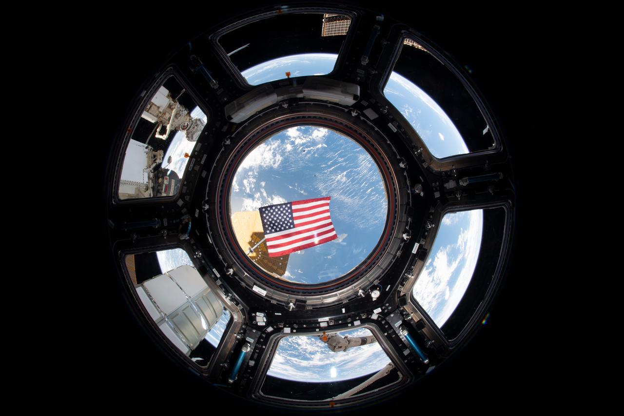 Photo of the American flag floating in front of all seven windows inside the Cupola during Expedition 68.
