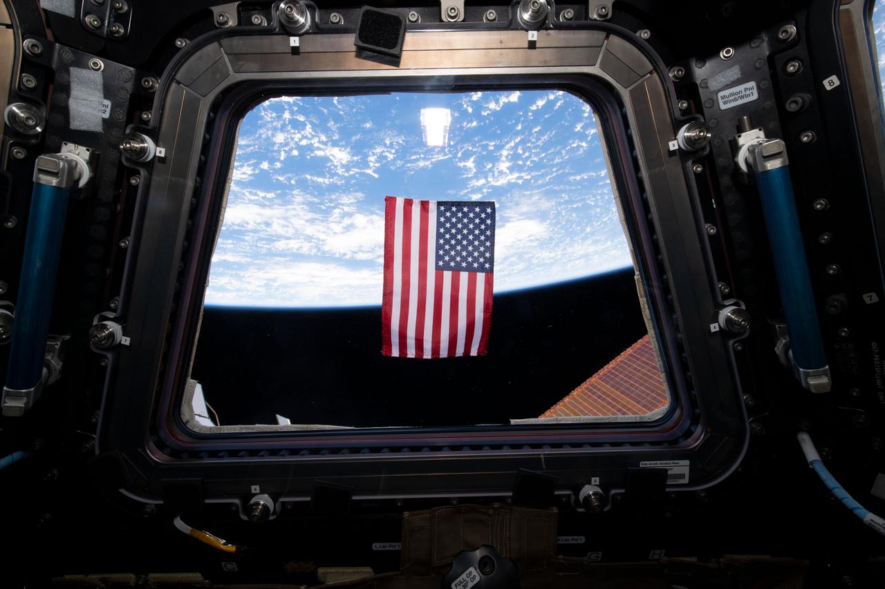 Photo taken during a crew activity with the American flag floating in front of the window in the Cupola during Expedition 68.