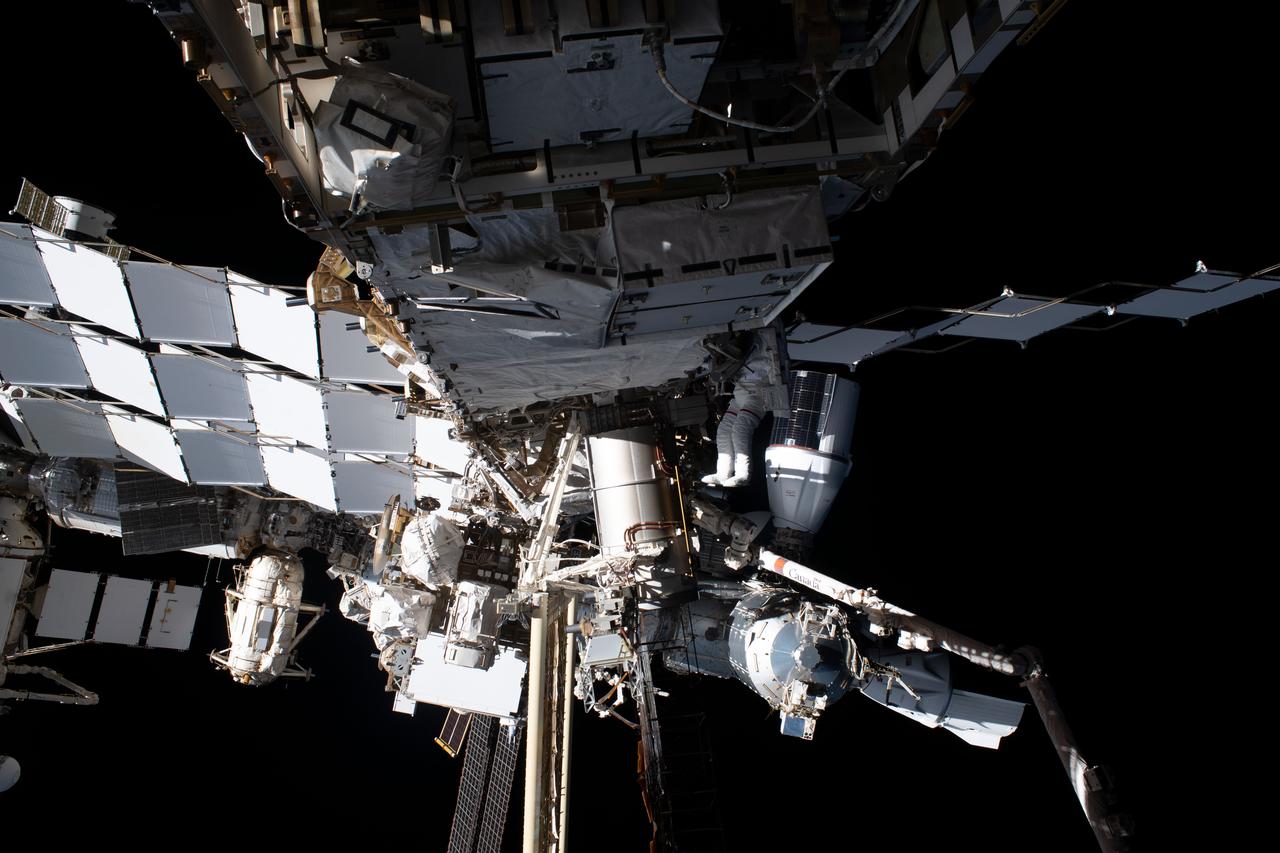 iss069e020815 (June 15, 2023) --- NASA astronaut Woody Hoburg is barely visible (center) tethered to the International Space Station's starboard truss structure during a spacewalk to install a roll-out solar array. Behind Hoburg, and at right, are the SpaceX Dragon cargo vehicle docked to the Harmony module's space-facing port and the SpaceX Dragon crew vehicle docked to Harmony's forward port.