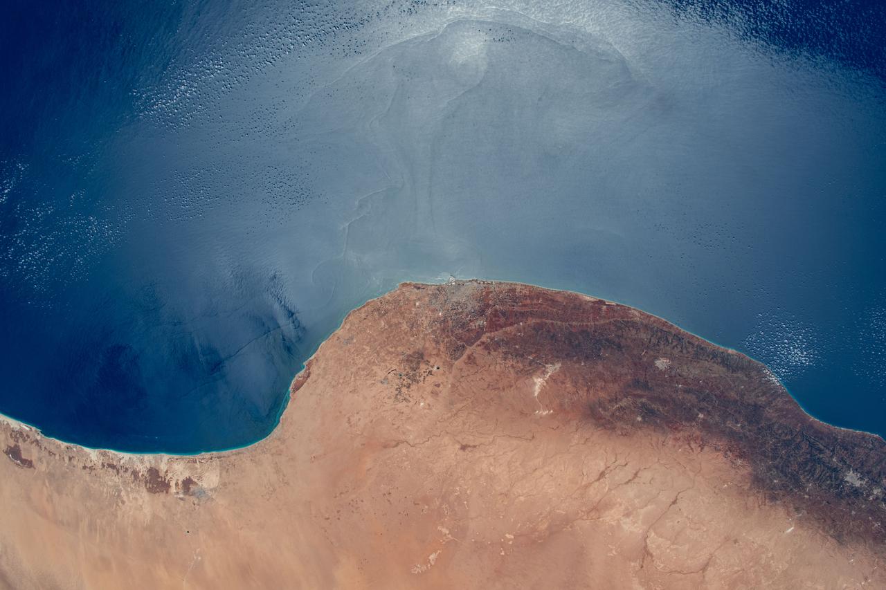 iss069e053975 (Aug. 4, 2023) --- The sun's glint and the deep blue waters of the Mediterranean Sea stand in stark contrast to the desert coast of Libya in this photograph from the International Space Station as it orbited 260 miles above. Benghazi, a city of 1.2 million people, is at center in the African nation's arid region in between its desert and temperate climates.
