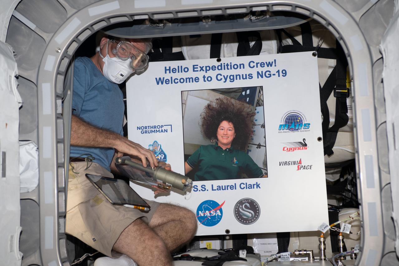 iss069e054159 (Aug. 4, 2023) --- NASA astronaut and Expedition 69 Flight Engineer Stephen Bowen wears personal protective equipment (PPE) shortly after entering the Northrop Grumman Cygnus space freighter. The PPE protects an astronaut from eye contact with and breathing in any dust or debris that may have dislodged during the spacecraft's ascent to orbit. Bowen poses with a portrait of NASA astronaut Laurel Clark, whom the Cygnus cargo craft is named after, who perished aboard space shuttle Columbia during its return to Earth on Feb. 1, 2003.
