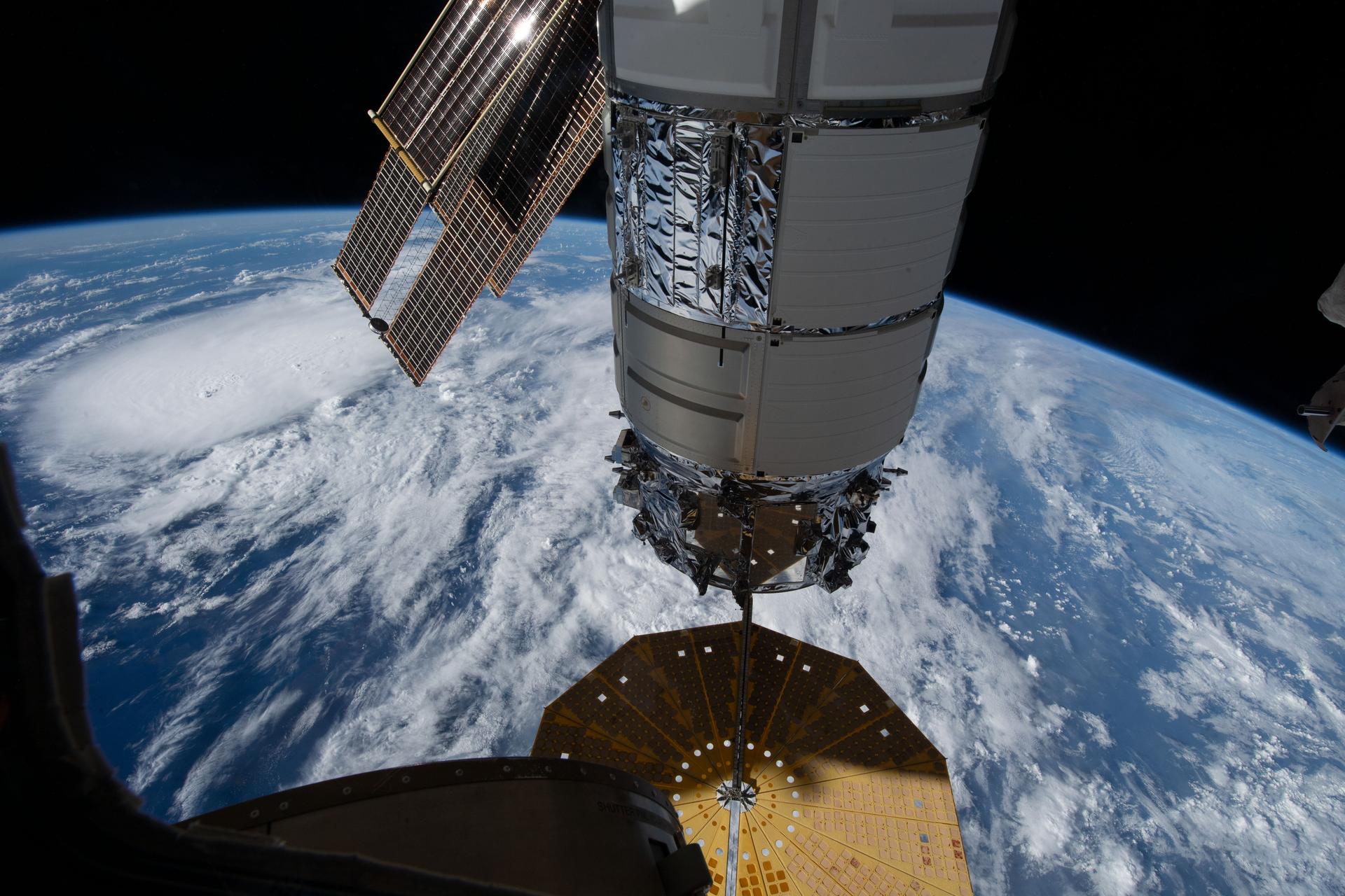 The cylindrical Cygnus space freighter, with white and silver panels and a large cymbal-shaped solar array at the bottom is attached to an Earth-facing port on the space station. A blue Earth with white clouds fills the background.