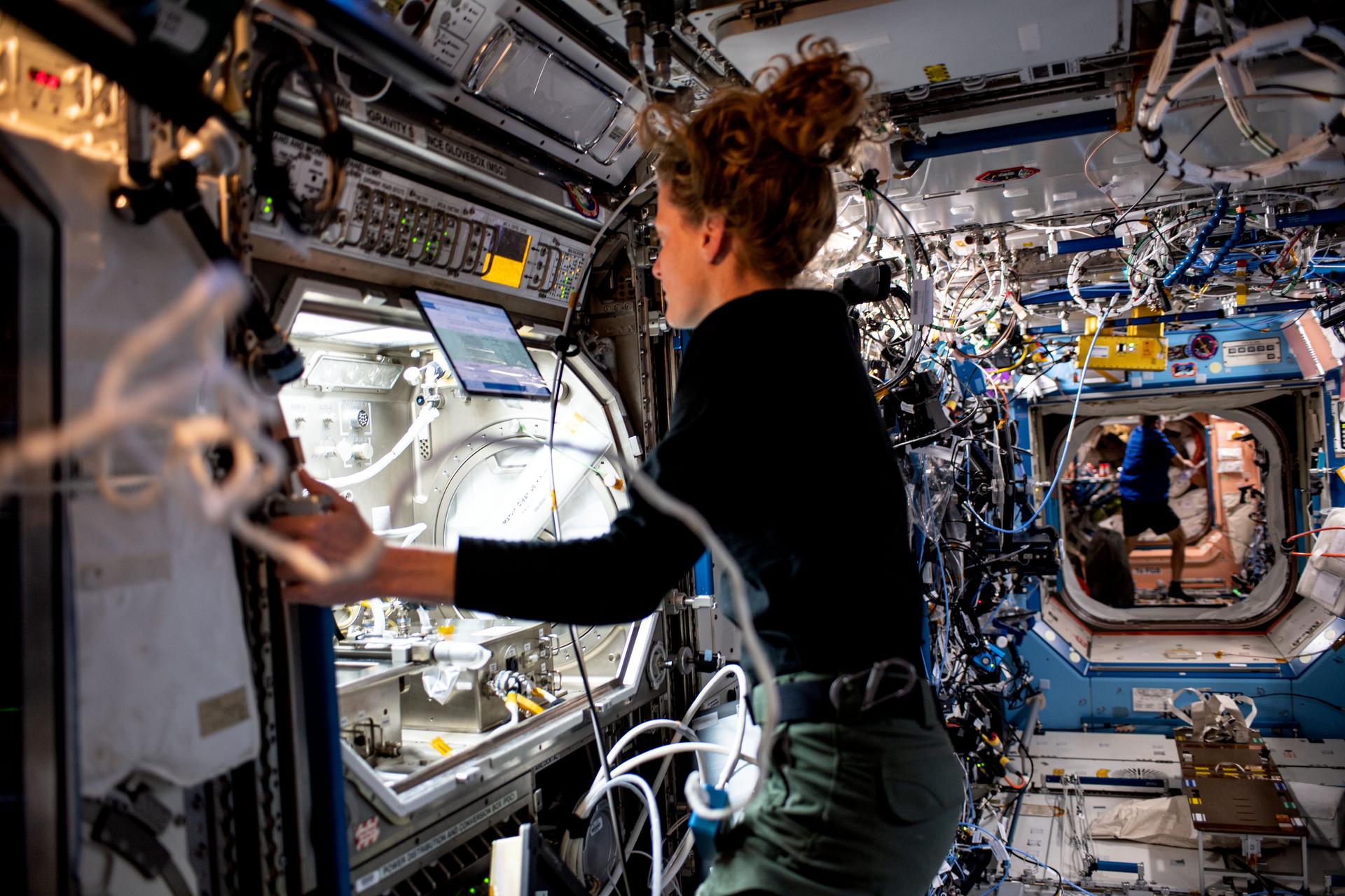 O'Hara is wearing a long-sleeved black shirt and green pants, with her hair in a ponytail. She is standing in front of the open front of a large, lighted module with two silver boxes inside it, each with multiple hoses and connectors. She is looking at a laptop screen at the top of the module and holding on to the edge of the hardware with her left hand.