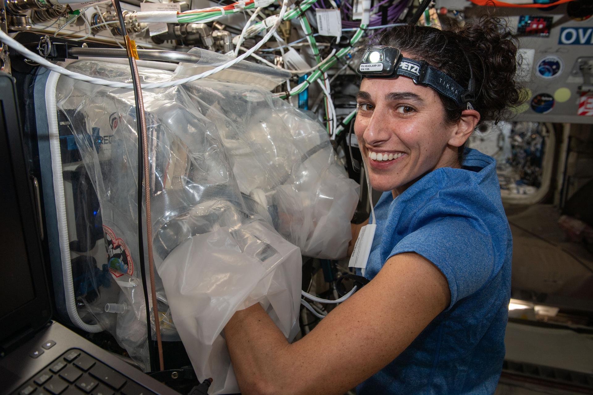 Jasmin Moghbeli smiles at the camera. She is wearing a short-sleeved blue shirt and a headlamp on her forehead and has both hands inside a plastic glovebag.