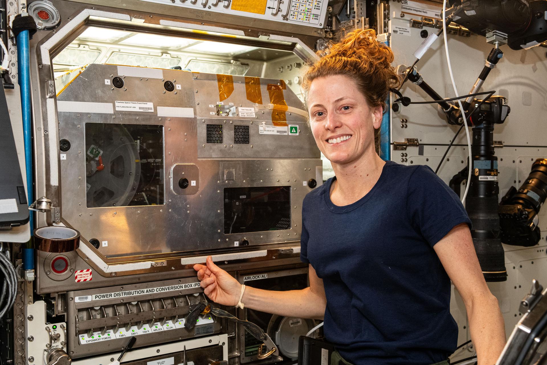 O'Hara, wearing a dark blue sleeveless t-shirt, smiles at the camera. Her right hand is touching the front of a large silver box built into a wall, with lights above it and a clear panel that reveals part of a circular sample holder that resembles an old film reel. There is a panel of switches below her hand and several large cameras on the wall behind her.