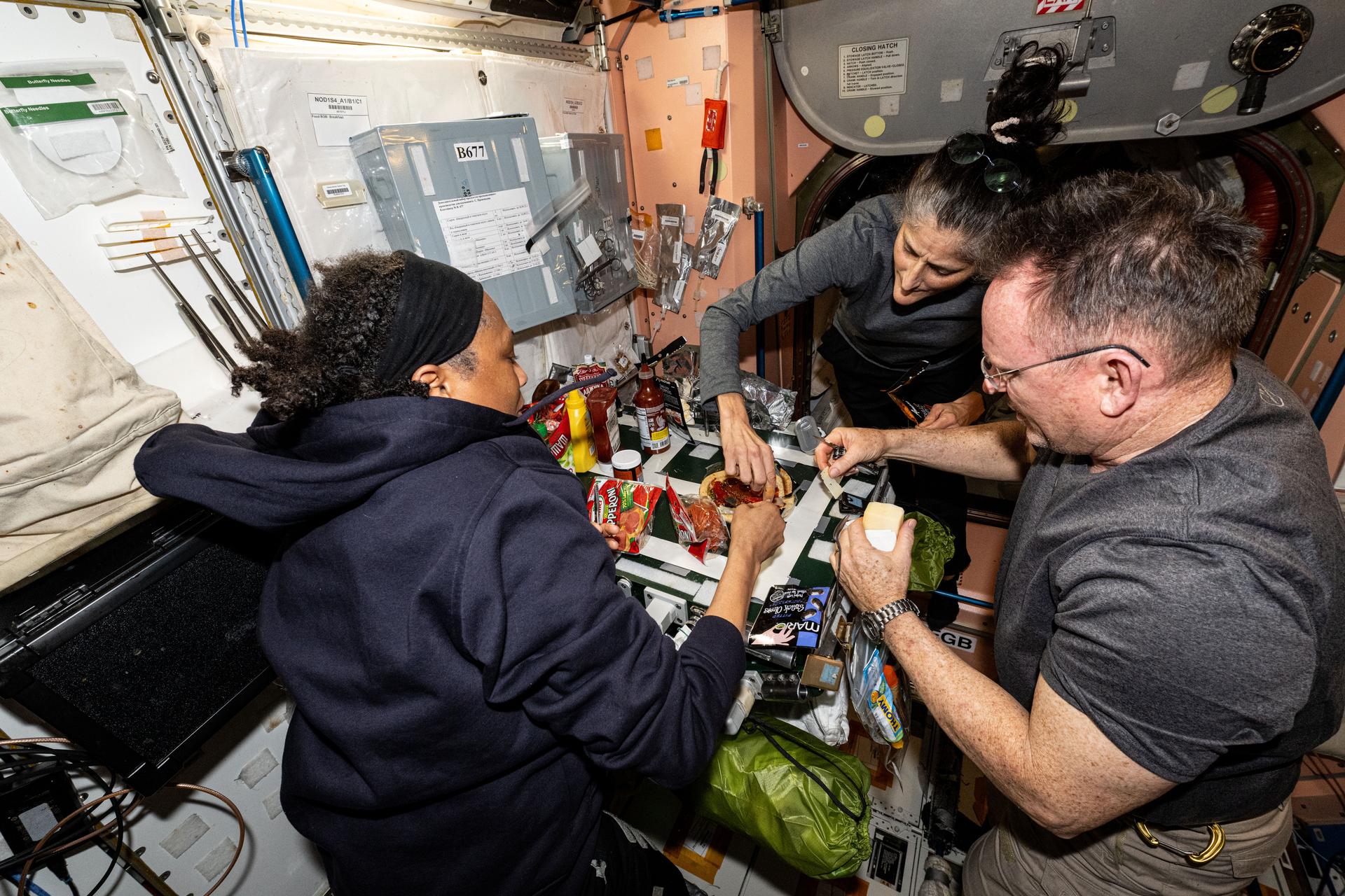 NASA astronauts (from left) Jeanette Epps, Suni Williams, and Butch Wilmore, all three Expedition 71 Flght Engineers, make pizza aboard the International Space Station's galley located inside the Unity module. Items are attached to the galley using tape and velcro to keep them from flying away in the microgravity environment.