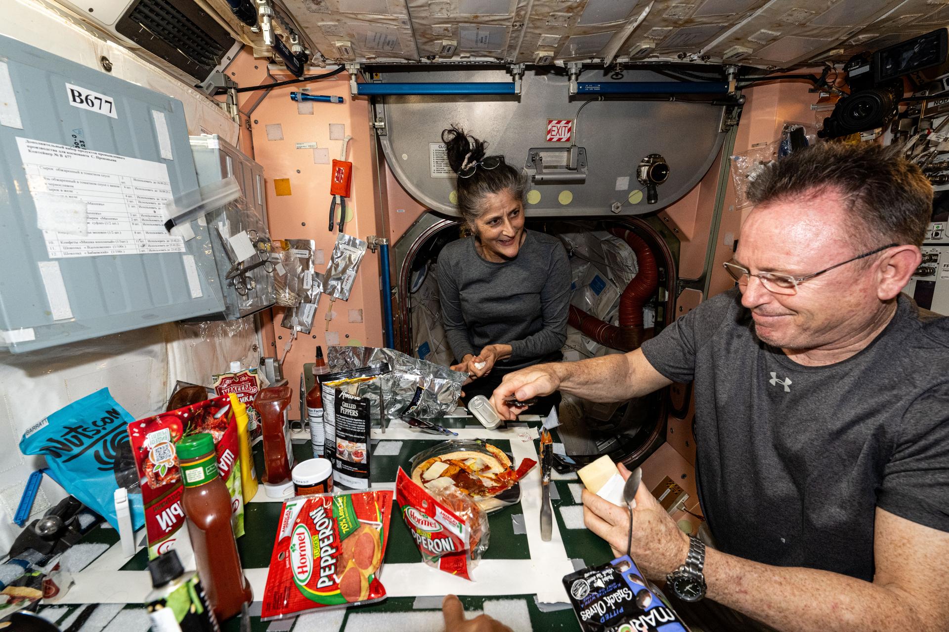 NASA astronauts Suni Williams and Butch Wilmore, both Expedition 71 Flght Engineers, make pizza aboard the International Space Station's galley located inside the Unity module. Items are attached to the galley using tape and velcro to keep them from flying away in the microgravity environment.