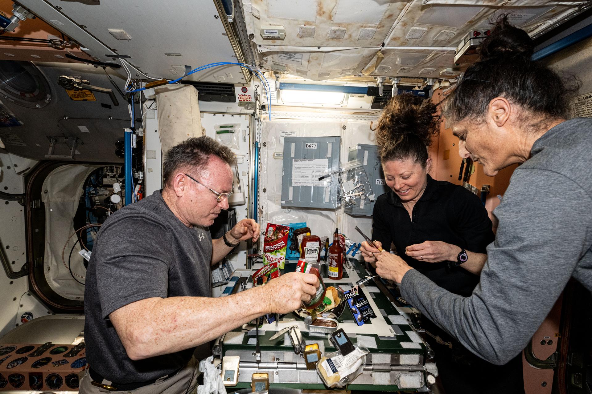 NASA astronauts (from left) Butch Wilmore, Tracy C. Dyson, and Suni Williams, all three Expedition 71 Flght Engineers, make pizza aboard the International Space Station's galley located inside the Unity module. Items are attached to the galley using tape and velcro to keep them from flying away in the microgravity environment.