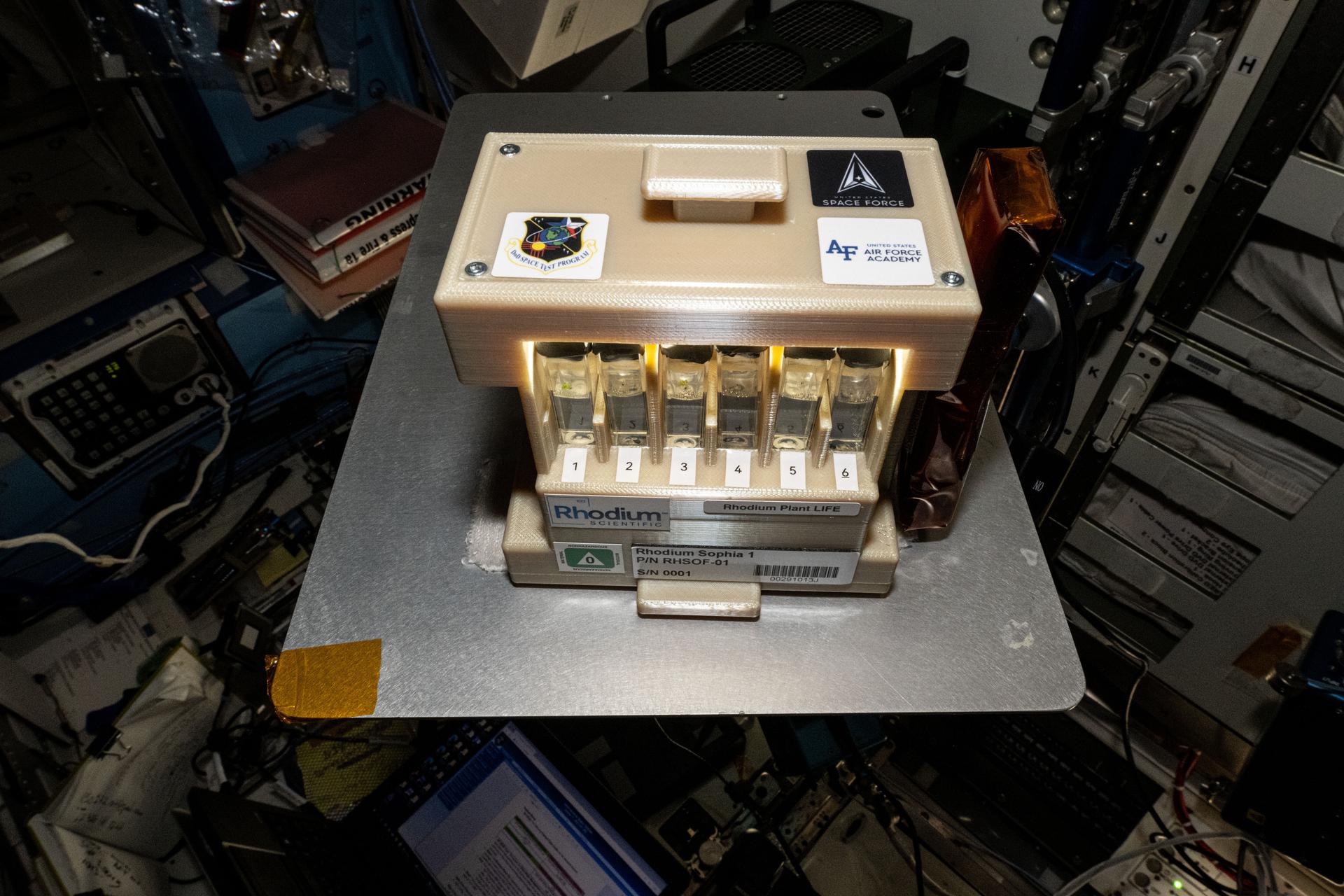 A tan box about the size of a small microwave sits on top of a silver metal tray. The box has stickers on the top that say, “DoD Space Test Program,” “United States Air Force Academy,” and “United States Space Force.” On its front are six numbered chambers holding vials filled with a clear liquid, and more labels below them, including one that says, “Rhodium Plant Life” and a barcode.